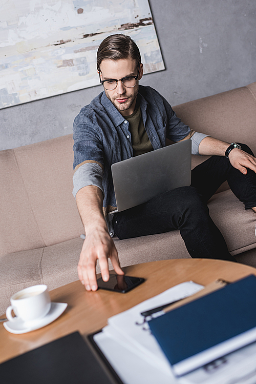 young handsome man with laptop reaching for smartphone