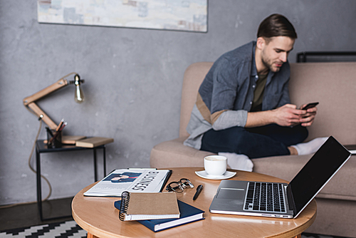 young handsome man using smartphone on couch with laptop and business supplies standing on table on foreground