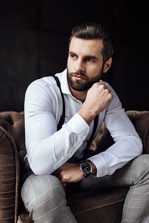 stylish man in suspenders thinking and sitting in armchair