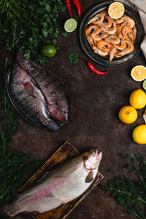 top view of raw fish, chili peppers, shrimp, herbs with lemons and tablecloth on dark table top