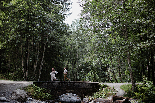young wedding couple running on bridge above mountain river in alpine forest