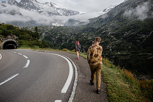 back view of man with smartphone photographing young woman walking on road near mountain tunnel in Alps