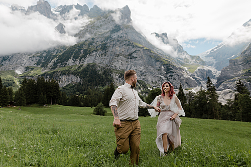 happy bride in wedding dress and groom holding hands and walking on alpine meadow with clouds