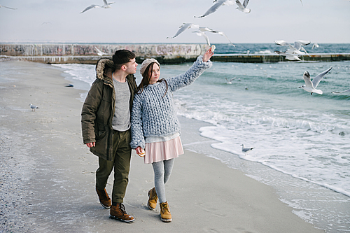 young couple walking on winter sea shore, girl pointing at seagulls