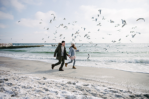 couple holding hands and running on winter sea shore with seagulls