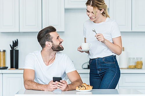 happy bearded man using smartphone and looking at smiling girlfriend holding cup of coffee at morning