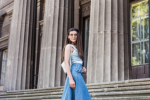 young stylish woman in eyeglasses and denim skirt standing on steps