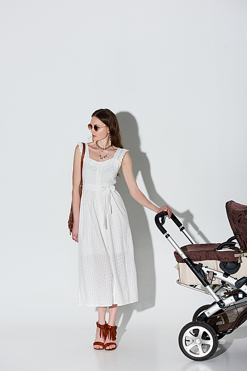 stylish woman in white dress and sunglasses standing with baby carriage and looking away on grey