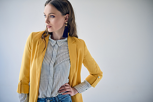 beautiful young woman in yellow jacket and striped shirt posing with hand on waist and looking away isolated on grey