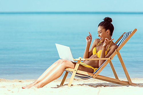 smiling african american woman gesturing by fingers and communicating by laptop while sitting on deck chair on sandy beach