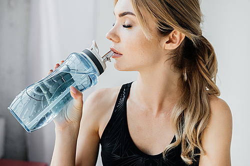 side view of athletic woman drinking water from sportive water bottle on grey backdrop