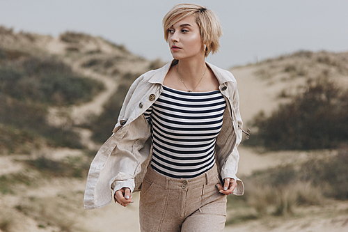 beautiful young woman in stylish striped shirt and vintage jacket walking on sand dunes