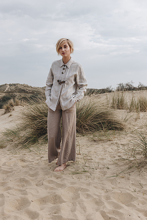 stylish young woman in retro jacket on sand dune under cloudy sky