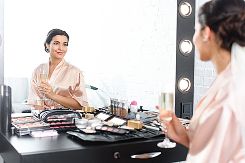 mirror reflection of smiling bride in silk bathrobe and veil with glass of champagne sitting at tabletop with cosmetics