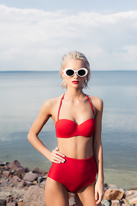 fashionable blonde 여성 posing in sunglasses and red bikini on rocky beach