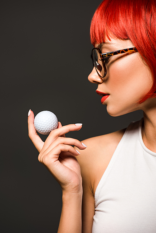 close-up portrait of beautiful young woman with red bob cut and stylish eyeglasses holding golf ball isolated on grey