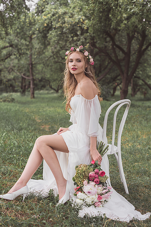 beautiful young bride with wedding bouquet sitting on chair and smiling at camera in park