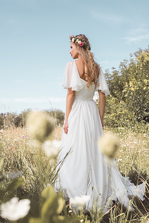 back view of beautiful young bride in wedding dress and floral wreath walking on field