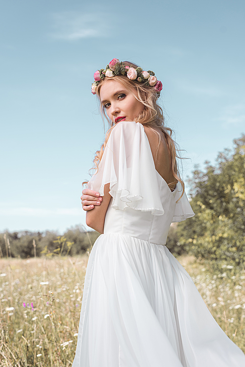 tender young bride in wedding dress and floral wreath standing on field and