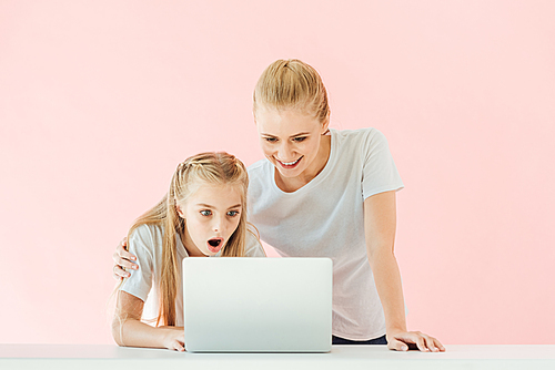 happy mother and surprised daughter in white t-shirts using laptop together isolated on pink