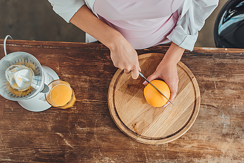 cropped image of woman cutting orange by knife on wooden board in kitchen