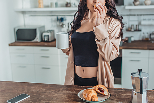 partial view of woman having breakfast with croissants and coffee at wooden table in kitchen at home