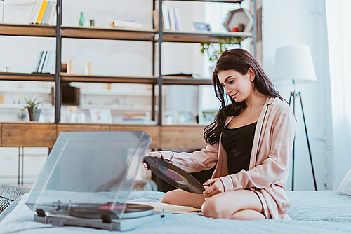 girl holding vinyl record near phonograph and sitting on bed at home