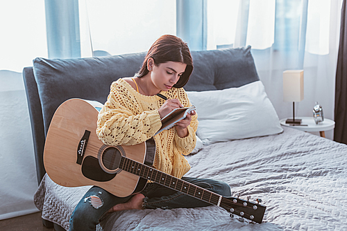beautiful girl sitting with acoustic guitar and writing song in textbook on bed at home