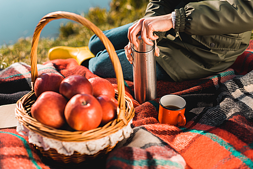cropped image of woman opening thermos while sitting on blanket with basket full of apples outdoors