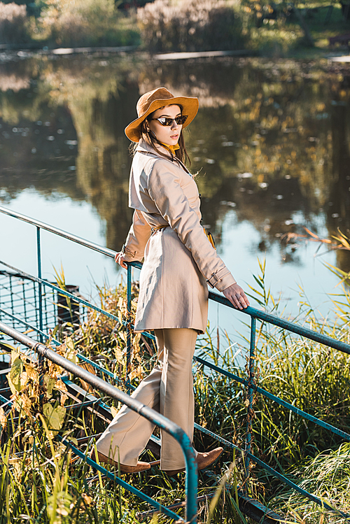 selective focus of stylish female model in sunglasses, trench coat and hat posing near pond in park