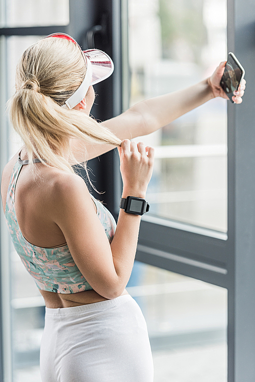 rear view of sportswoman with smartwatch taking selfie on smartphone at gym