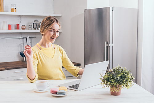 happy woman holding credit card while looking at laptop in kitchen