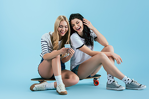 Laughing girls sitting on longboard with smartphone on blue background