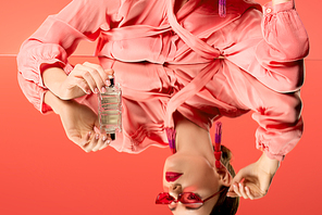 cropped view of woman in blouse and red sunglasses posing with perfume bottle and mirror reflection isolated on living coral