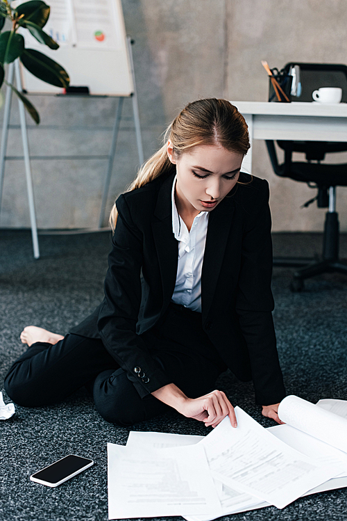 barefoot businesswoman sitting on floor near work-table and reading documents