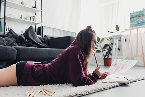 beautiful designer lying on carpet, biting pencil and looking at sketch in living room