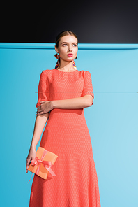 fashionable woman in living coral dress holding gift box on blue