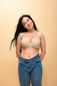 smiling young woman in blue jeans and bra holding hands behind back while  isolated on beige, body positivity concept