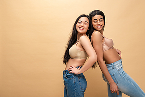 two pretty multicultural girls in blue jeans and bra standing back to back and smiling isolated on beige, body positivity concept