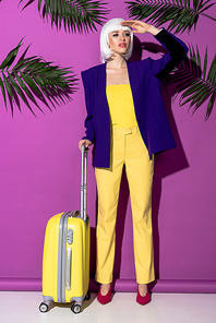 Stylish woman in wig with suitcase looking away on purple background