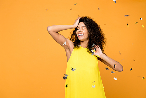 cheerful curly african american woman smiling near shiny confetti stars on orange