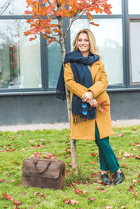 beautiful stylish woman in yellow coat and scarf standing at tree in autumn park