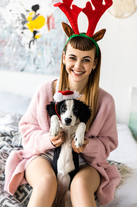 portrait of smiling woman with deer horns on head and puppy in santa hat