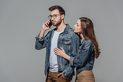 young woman touching and looking at stylish man in eyeglasses talking on smartphone isolated on grey