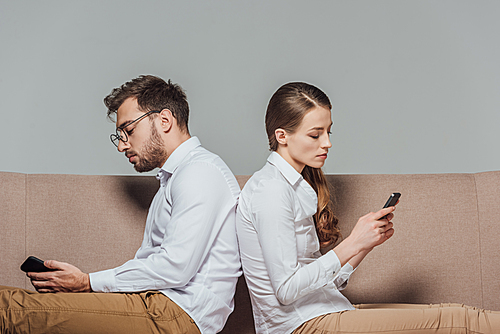 young couple sitting back to back and using smartphones isolated on grey