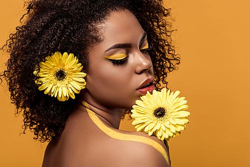 Attractive young american woman with artistic make-up smelling gerbera isolated on orange background