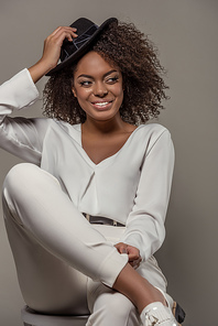 Beautiful african american woman in white clothes smiling and holding black hat isolated on grey background