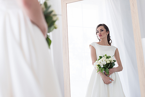 young bride in dress with wedding bouquet looking at her reflection in mirror