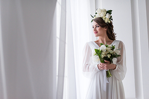 attractive bride in elegant dress and floral wreath holding wedding bouquet