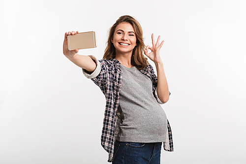 happy young pregnant woman taking selfie with smartphone and showing ok sign isolated on white